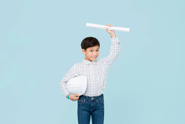 Cute smiling young Asian boy with helmet aspiring to be future engineer showing blueprint isolated on light blue studio background - Foto, Imagen
