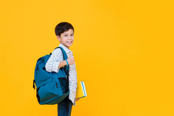 Smiling handsome mixed race schoolboy with books and backpack giving thumbs up isolated on yellow background with copy space - Zdjęcie, obraz