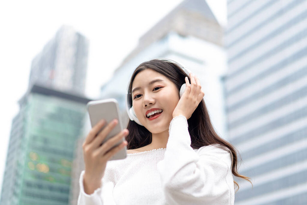 Happy uśmiechnięta Azjatka w słuchawkach i patrząc na telefon komórkowy podczas słuchania strumieniowej muzyki na tle budynku miasta - Zdjęcie, obraz