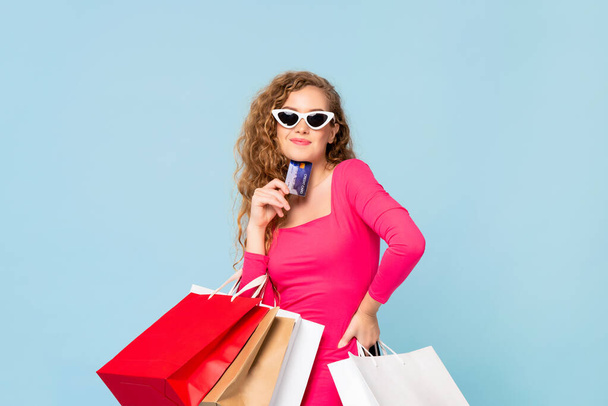 Elinde alışveriş poşetleri olan mutlu, güzel, beyaz bir kadın mavi arka planda kredi kartı gösteriyor. - Fotoğraf, Görsel