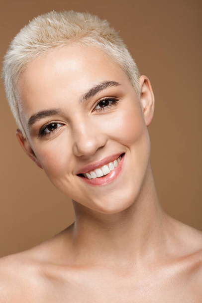 Afbeelding van een mooie vrolijke optimistische glimlachende jonge blonde stijlvolle vrouw met kort kapsel poseren geïsoleerd over donker beige muur achtergrond. - Foto, afbeelding