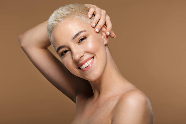 Afbeelding van een vrij vrolijke glimlachende jonge blonde stijlvolle vrouw met kort kapsel poseren geïsoleerd over donker beige muur achtergrond. - Foto, afbeelding