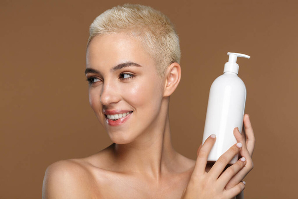 Image of a pretty cheerful smiling young blonde stylish woman with short haircut posing isolated over dark beige wall background holding bottle with cream lotion. - Fotoğraf, Görsel