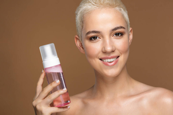 Image of a beautiful happy optimistic smiling young blonde stylish woman with short haircut posing isolated over dark beige wall background holding bottle with cosmetics water. - Foto, afbeelding