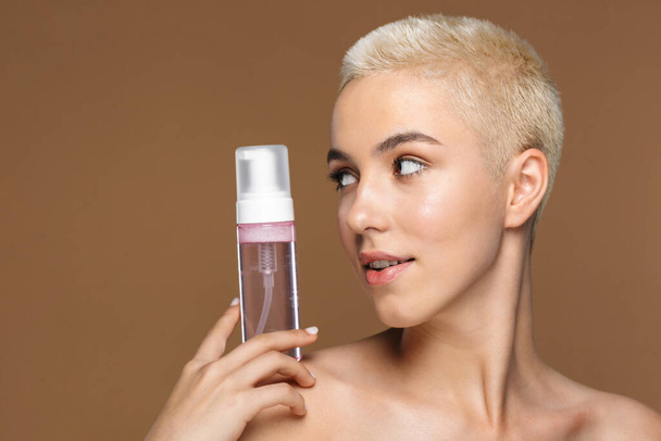 Image of young pretty blonde stylish woman with short haircut posing isolated over dark beige wall background holding bottle with cosmetics water. - Φωτογραφία, εικόνα