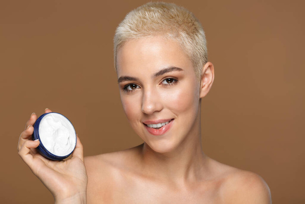 Image of a beautiful happy optimistic smiling young blonde stylish woman with short haircut posing isolated over dark beige wall background holding cream. - Zdjęcie, obraz