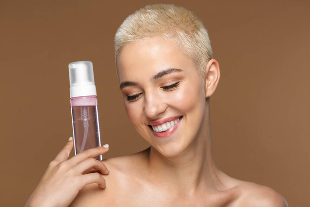 Image of a pretty cheerful smiling young blonde stylish woman with short haircut posing isolated over dark beige wall background holding bottle with cosmetics water. - Foto, Imagem