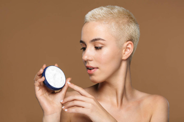 Picture of a beautiful young blonde stylish woman with short haircut posing isolated over dark beige wall background holding cream. - Foto, Imagem