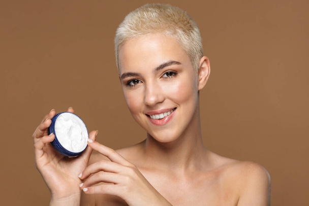 Image of a pretty cheerful smiling young blonde stylish woman with short haircut posing isolated over dark beige wall background holding cream. - Foto, Imagem
