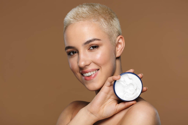 Afbeelding van een mooie vrolijke optimistische glimlachende jonge blonde stijlvolle vrouw met kort kapsel poseren geïsoleerd over donker beige muur achtergrond holding crème. - Foto, afbeelding