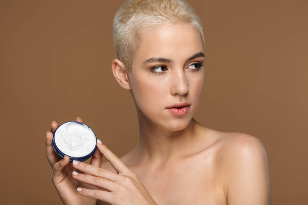 Picture of a beautiful young blonde stylish woman with short haircut posing isolated over dark beige wall background holding cream. - 写真・画像