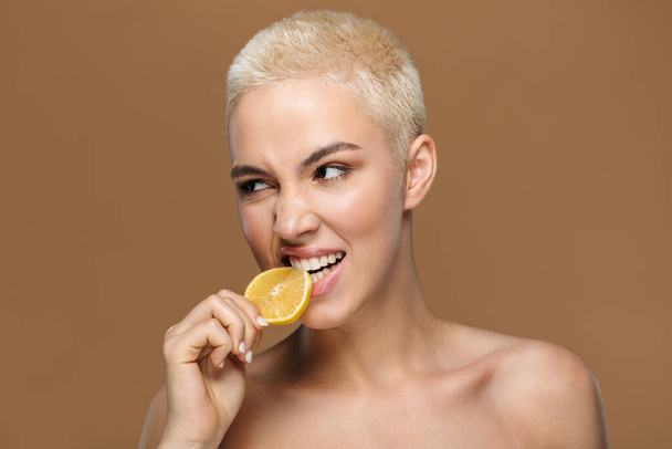 Image of a beautiful young blonde stylish woman with short haircut posing isolated over dark beige wall background bite lemon. - Valokuva, kuva