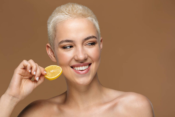 Image of a beautiful happy optimistic smiling young blonde stylish woman with short haircut posing isolated over dark beige wall background with lemon. - Fotoğraf, Görsel