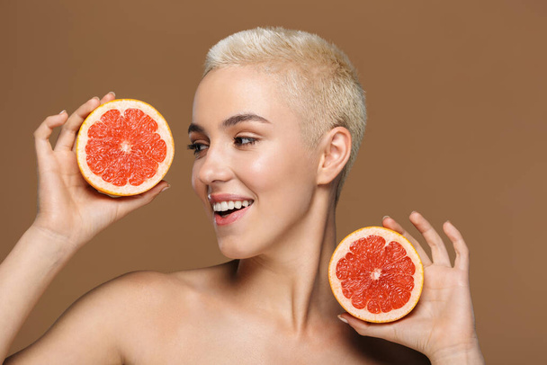 Image of a pretty cheerful smiling young blonde stylish woman with short haircut posing isolated over dark beige wall background holding grapefruit. - Photo, Image
