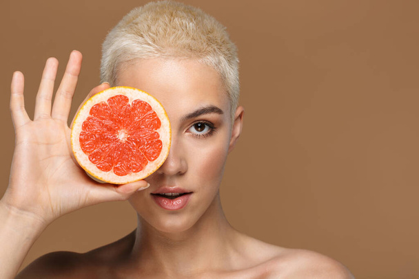 Beauty portrait of an attractive young topless blonde short haired woman holding grapefruit isolated over beige background - Foto, imagen