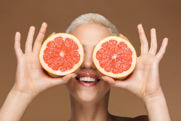 Image of a beautiful happy optimistic smiling young blonde stylish woman with short haircut posing isolated over dark beige wall background holding grapefruit covering eyes. - Fotó, kép