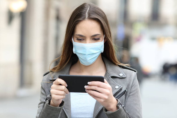 Serious woman with protective mask avoiding contagion watching video online on smart phone in the street - Foto, Imagen