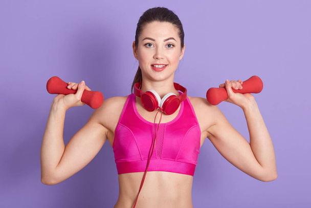 Image of cheerful inspired young female standing isolated over lilac background in studio, looking directly at camera, raising dumbbells in hands, keeping fit, having headphones around neck. - 写真・画像