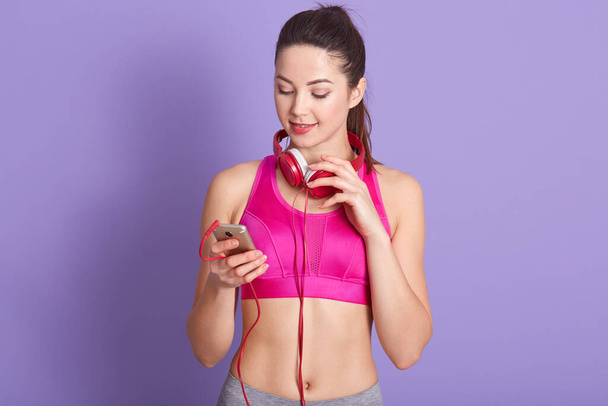 Studio shot of attractive woman wearing sports wear, having headphones around neck, holding smart phone in hands, choosing music for workout, sportswoman being ready to train in gym. Fitness concept. - Foto, afbeelding