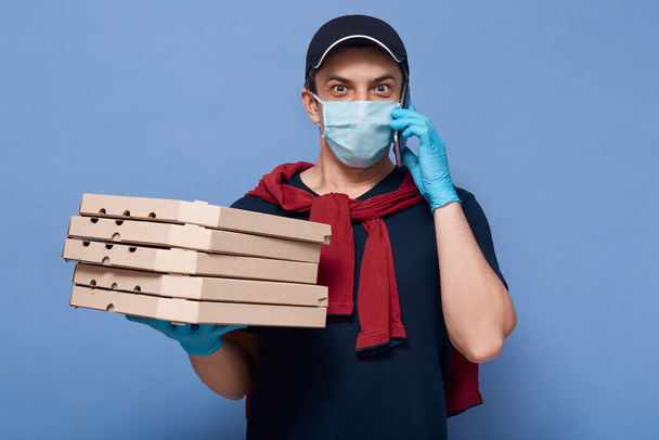 Studio shot of astonished courier talking via smart phone with her customer, has surprised facial expression, holding stack of pizzas, wearing casual clothes and protective face mask and gloves. - Photo, image