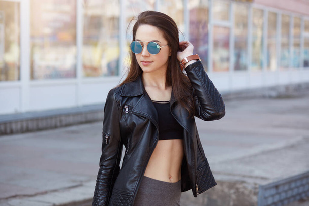 Closeup portrait of adorable confident female posing outdoors, wearing stylish clothing and sunglasses, looking in distance, standing with bared belly, keeps hand on her hair, expresses confidence. - Photo, image