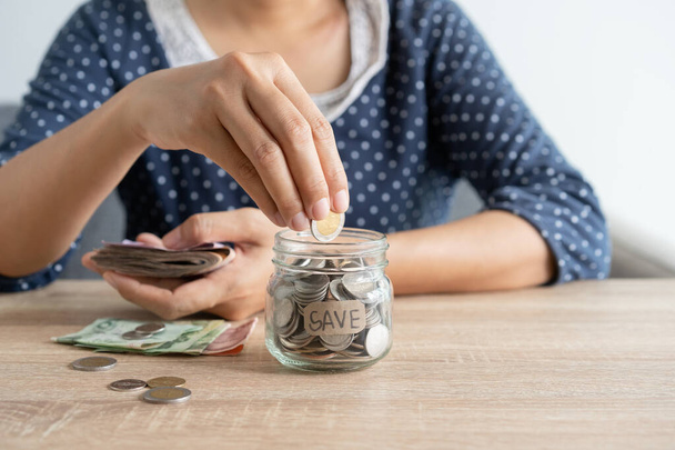 Asian woman hand is putting a coin in a glass bottle and a pile of coins on the table at home, Investment business and retirement. finance and saving money for the future concept - Photo, image