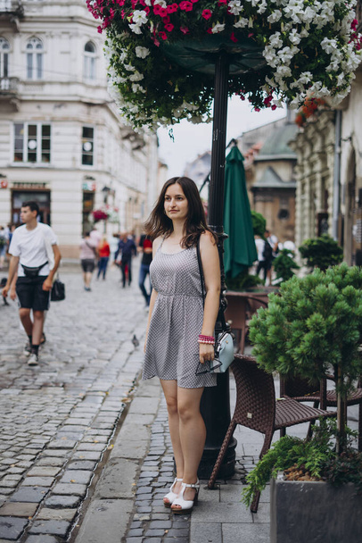 Young woman in summer dress downtown on flowers background. Woman in old town. The girl is walking in a beautiful old city in a gray dress. - Photo, Image