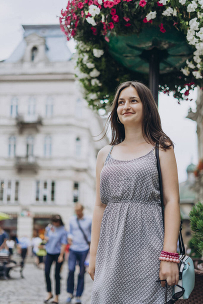 Beautiful girl walking in the summer city. Girl in a summer dress on a background of flowers. Young woman in a dress and with a handbag walks downtown. - Photo, Image