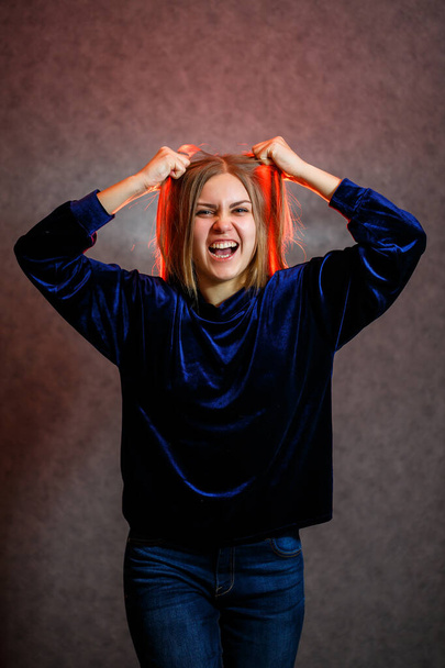 Chica con el pelo rubio posando emocionalmente sobre un fondo gris. Su cabello está iluminado en rojo - Foto, Imagen