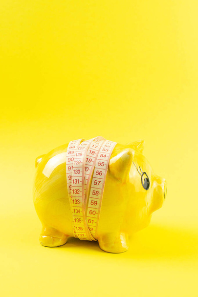 Gele spaarpot in de vorm van een varken gewikkeld met meetlint op een gele achtergrond. Het concept van geld besparen om gewicht te verliezen. - Foto, afbeelding