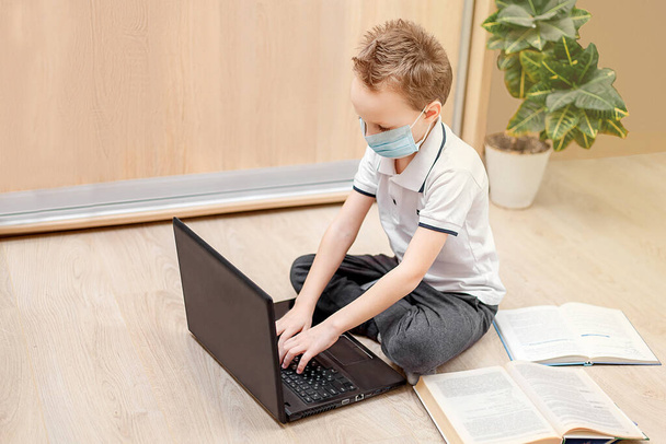 Evde öğrenim çevrimiçi. Koruyucu maskeli bir çocuk yerde oturur ve bilgisayarda çalışır. Okul çocuğu ev ödevi yapıyor. Karantinaya alın. Coronavirus. Evde kal. Evde eğitim. Kendine dikkat et.. - Fotoğraf, Görsel