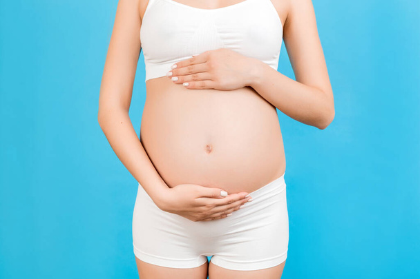Karnına sarılan hamile bir kadın görüntüsü. Annem mavi arka planda beyaz iç çamaşırı giyiyor. Müstakbel ebeveyn çocuğunu bekliyor. Boşluğu kopyala. - Fotoğraf, Görsel