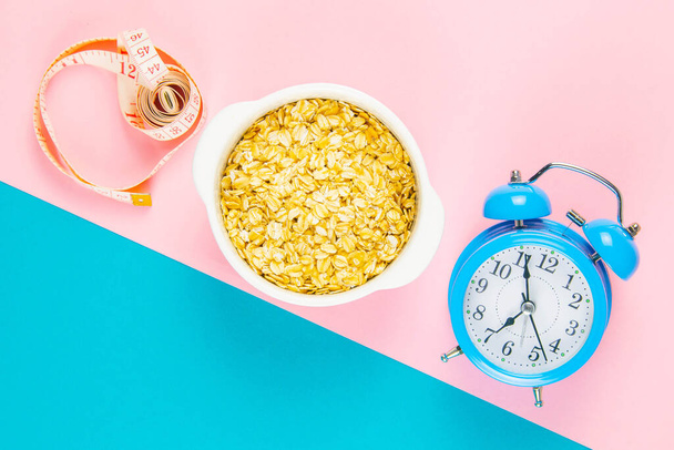 Blue alarm clock with oatmeal and measuring tape on a pink and blue background. Healthy nutrition concept. Top views with clear space. - Фото, изображение