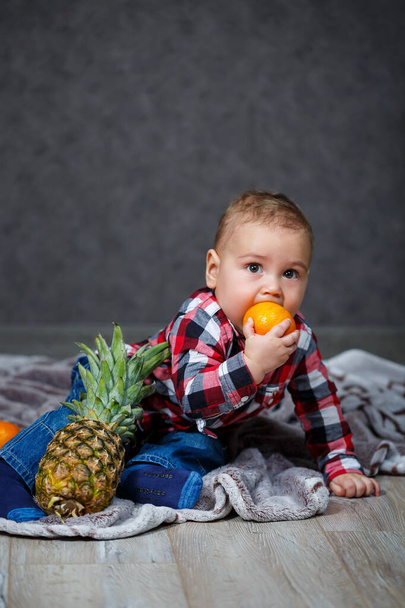 El niño en la camisa se sienta en el cuadros y sostiene la fruta
 - Foto, Imagen