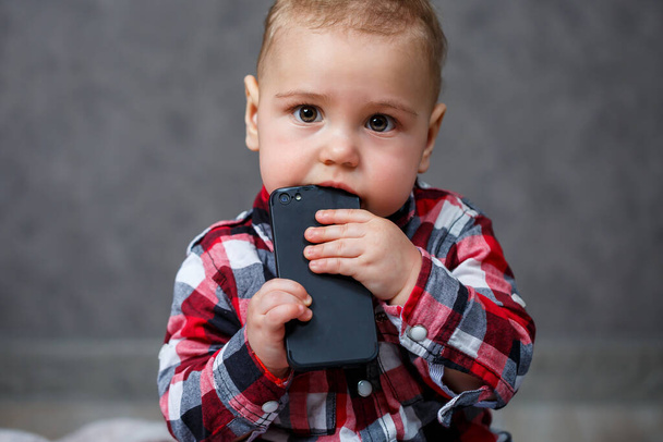 Μικρό αγόρι με πουκάμισο ροκανίζει το κινητό τηλέφωνο - Φωτογραφία, εικόνα