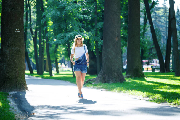young beautiful girl in denim overalls and a light hat walking in the park - Photo, Image