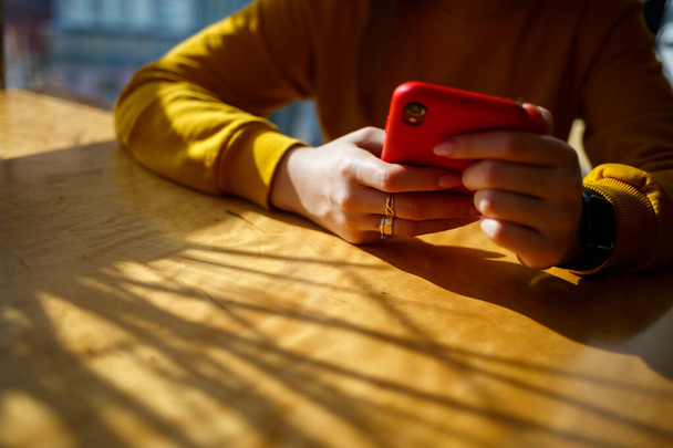 lány egy sárga pulóverben ül egy nagy ablak mellett egy piros telefonnal - Fotó, kép
