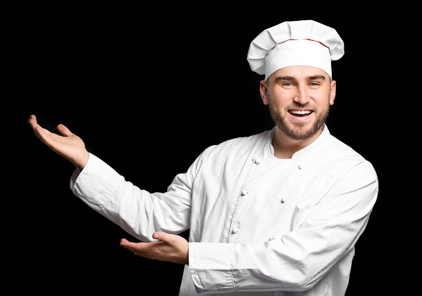 Knappe mannelijke chef-kok toont iets op donkere achtergrond - Foto, afbeelding