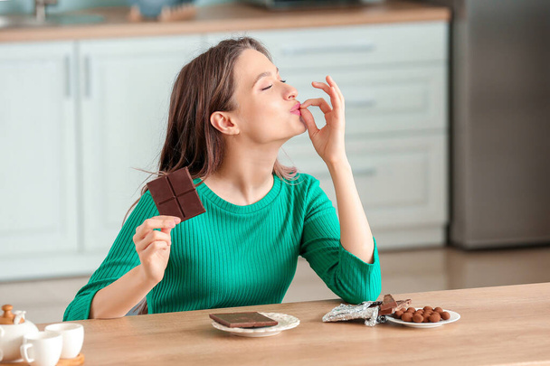 Beautiful young woman eating chocolate in kitchen - Zdjęcie, obraz