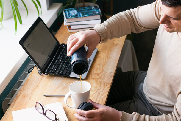 Молодой человек наливает горячий кофе или чай, работает дома на цифровом ноутбуке, сидит за столом. Концепция дистанционной работы
 - Фото, изображение