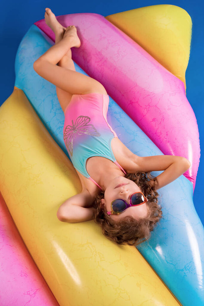 Hezké usmívající se dívka na sobě růžové a modré plavky a sluneční brýle leží, relaxovat na duhové nafukovací matrace. - Fotografie, Obrázek
