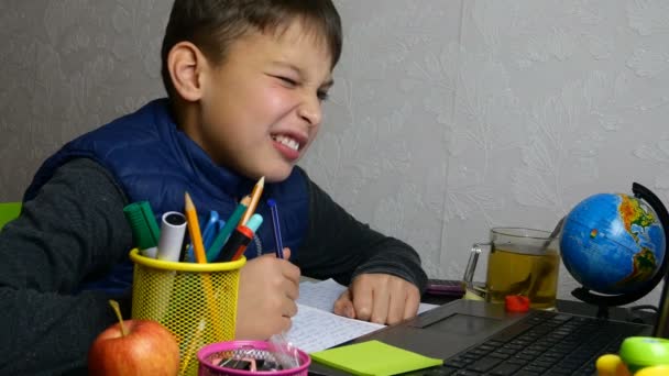 Távolságtanulás. Vicces iskolás fiú online tanul, és iskolai házi feladatot csinál a jegyzetfüzetével karantén alatt. Grimaszol és bolondozik. Közelkép portré. - Felvétel, videó