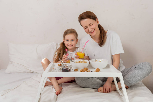 Happy mom and daughter have healthy breakfast on bed in a light bedroom on a sunny morning. They drink orange juice through a straw and laugh. Healthy food and family relationships concept. Good mood. - Photo, image