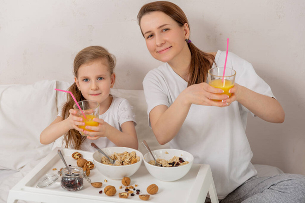 Happy mom and daughter have healthy breakfast on bed in a light bedroom on a sunny morning. They hold glasses with orange juice and smile. Healthy food and family relationships concept. Good mood. - Foto, imagen