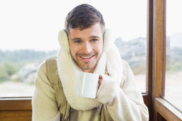 Smiling man wearing earmuff with coffee cup against window - Foto, afbeelding