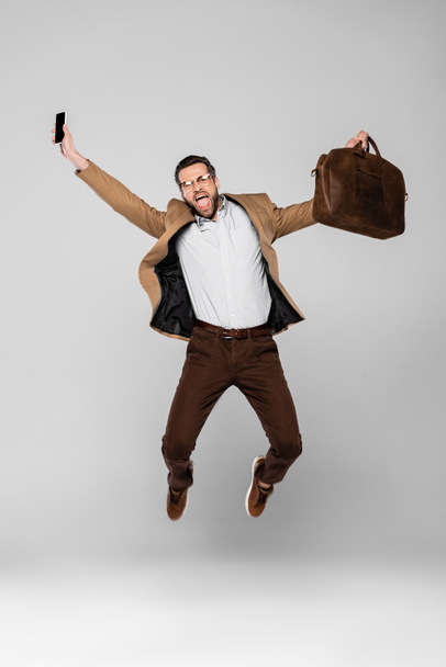 excited businessman holding smartphone with blank screen and bag while levitating on grey  - Φωτογραφία, εικόνα