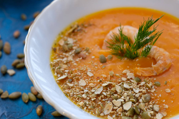 close-up pumpkin soup with prawns, pumpkin seeds whole and broken and decorated with dill in a ligth blue plate on darkk blue background. Horizontal top view. - Zdjęcie, obraz