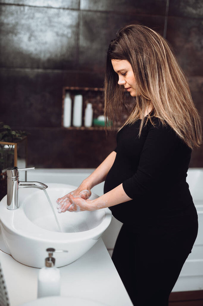 Zwangere vrouw glimlachend en handen wassen thuis met behulp van ontsmettingsmiddel en zeep, leidingwater en schoonmaken van cosmetica - Foto, afbeelding