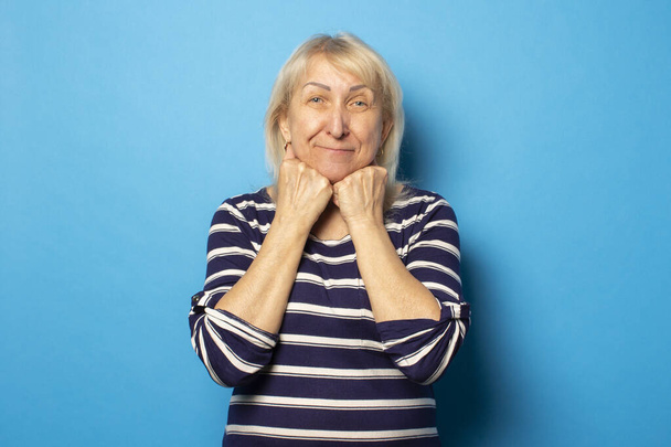 Portré egy öreg, barátságos nőről alkalmi pólóban, aki az állán tartja a kezét egy elszigetelt kék háttéren. Érzelmi arc. - Fotó, kép