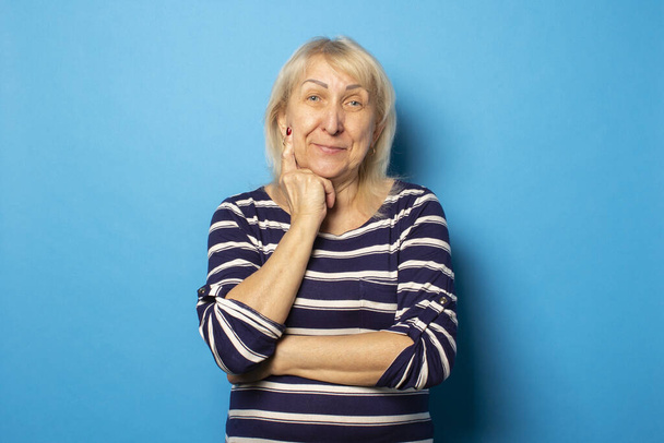 Портрет старої дружньої жінки в повсякденній футболці тримає її руки на підборідді на ізольованому синьому фоні. Емоційне обличчя
. - Фото, зображення